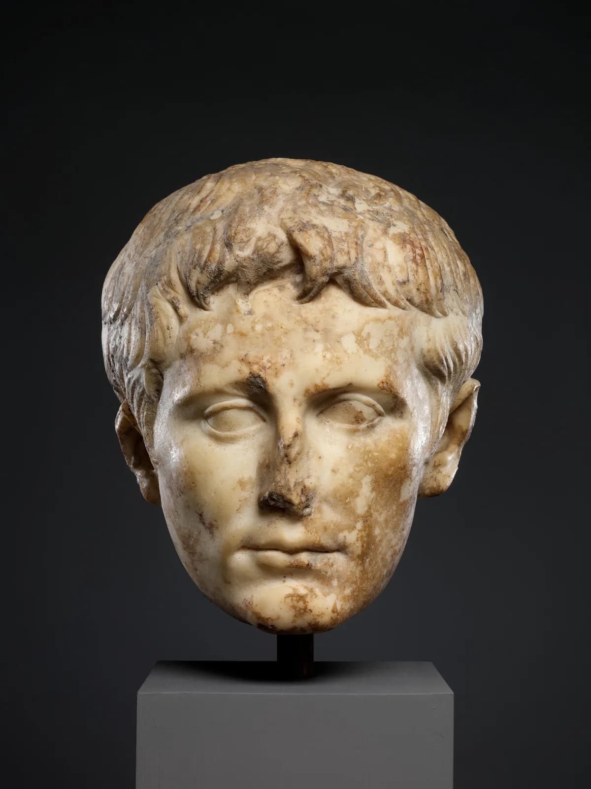 A Roman marble portrait head of the emperor Augustus on a plain grey plinth against a black background: young, clean-shaven, hair combed forward in flat locks, the stone stained brown across one cheek and the nose broken away