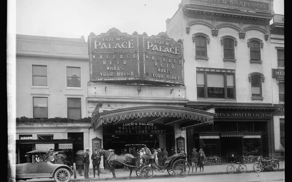 The exterior of Loew's Palace Theater in 1920, with an illuminated marquee and period cars and carriages outside