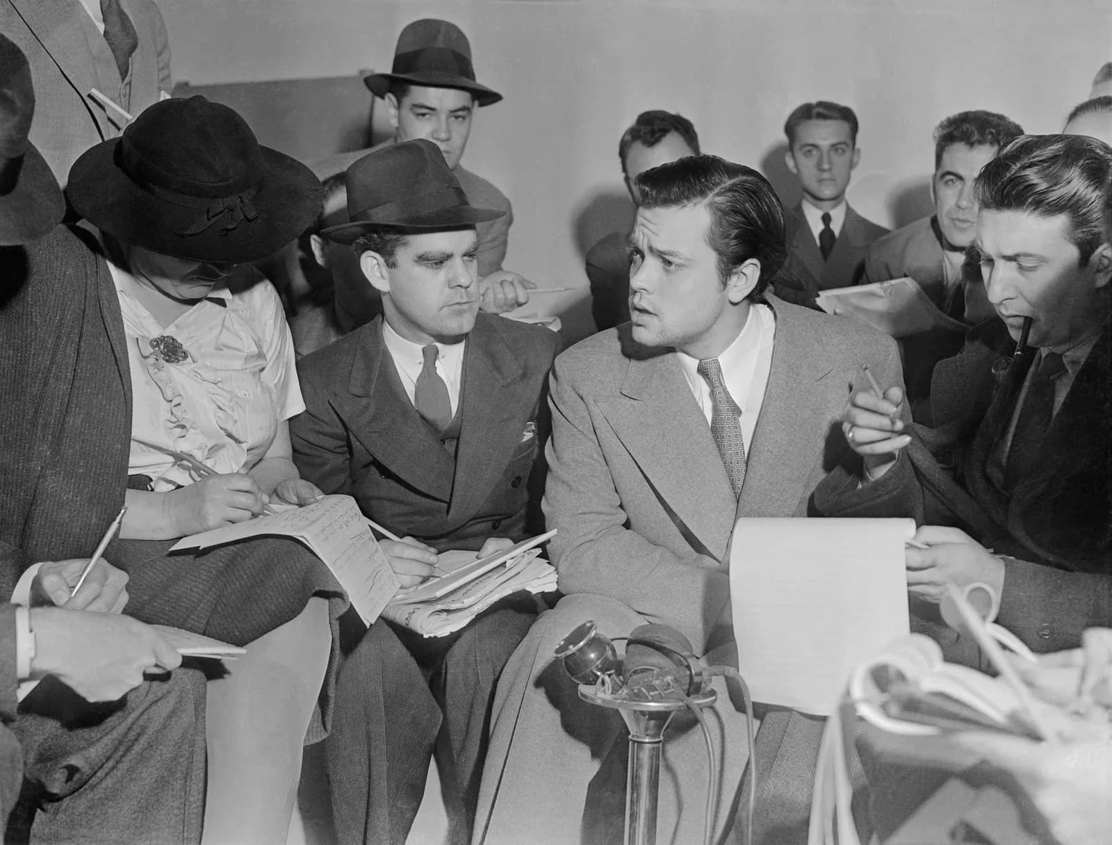 A black and white press photograph of Orson Welles seated among reporters in hats, leaning forward to speak while they take notes and hold microphones