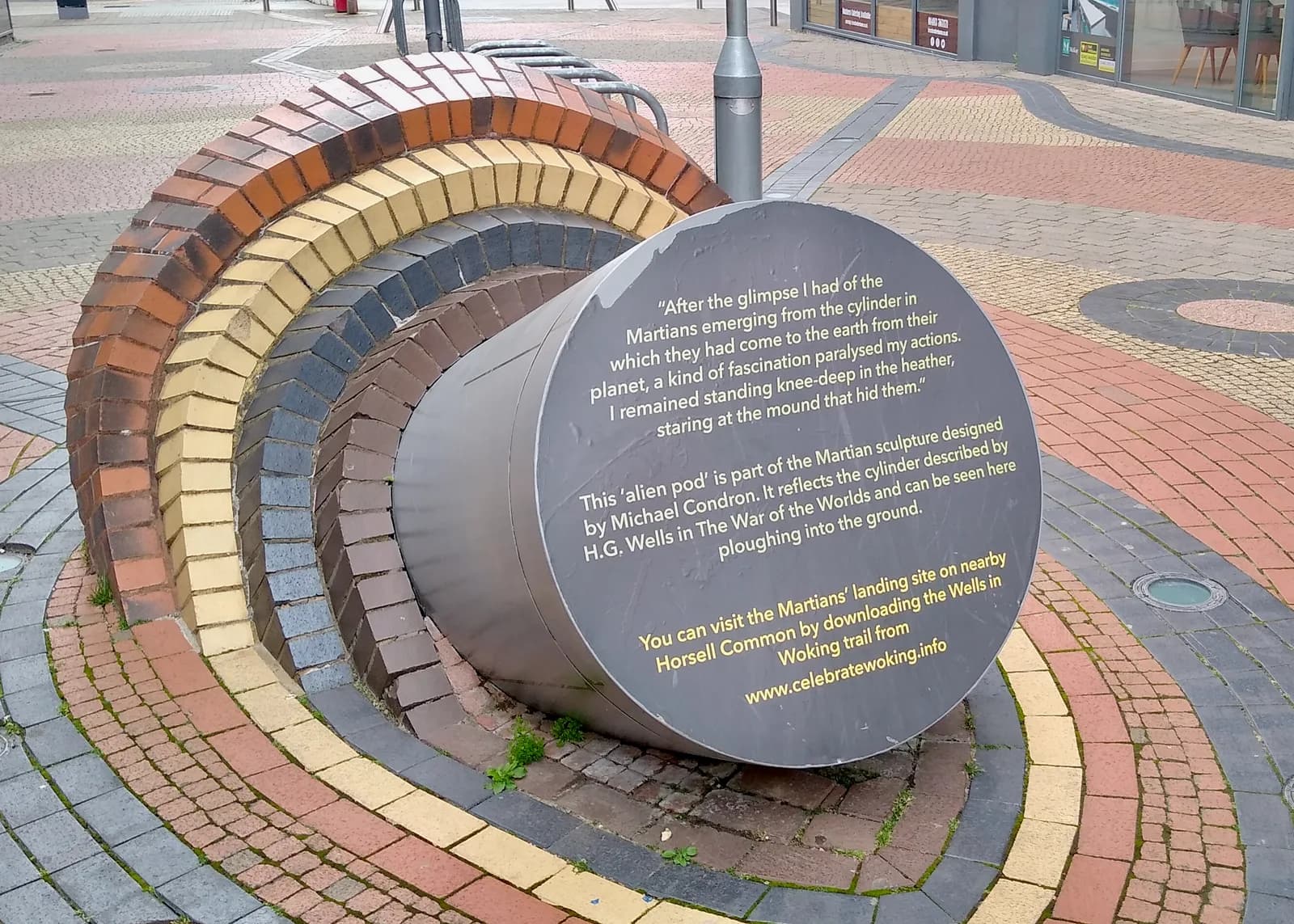 A steel sculpture of a Martian cylinder half buried in a paved town square, ringed by coloured paving stones that ripple outwards from it, with an explanatory plaque on the end