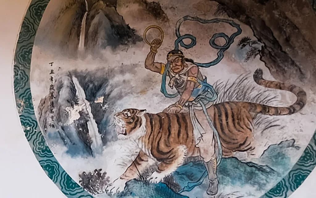 A circular temple wall mural showing Wu Song astride a tiger on a rocky slope, one arm raised, with a waterfall behind him